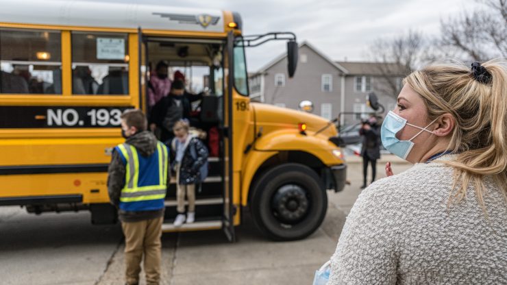 An educator with long blonde hair in a ponytail and cream-beige sweater wears a mask and looks off to the left. She stands near a yellow school bus. A student with a blue jacket and backpack stands on the steps of the bus while a crossing guard in a neon vest stands nearby.