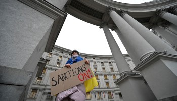 A protester wearing a Ukraine flag over her shoulders holds a sign that reads "Sanctions on!"