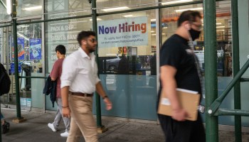 A "now hiring" sign is displayed in a window in Manhattan, New York. Several people are walking past it on the sidewalk.
