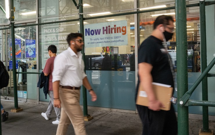 A "now hiring" sign is displayed in a window in Manhattan, New York. Several people are walking past it on the sidewalk.