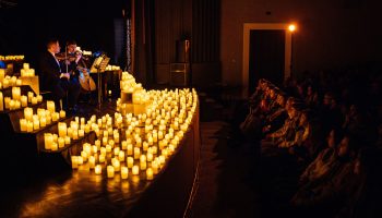 On a dark stage, two people in suits play a cello and violin or viola surrounded by dozens of candles. To the right is the audience, rows and rows of people whose faces are lit by candlelight.