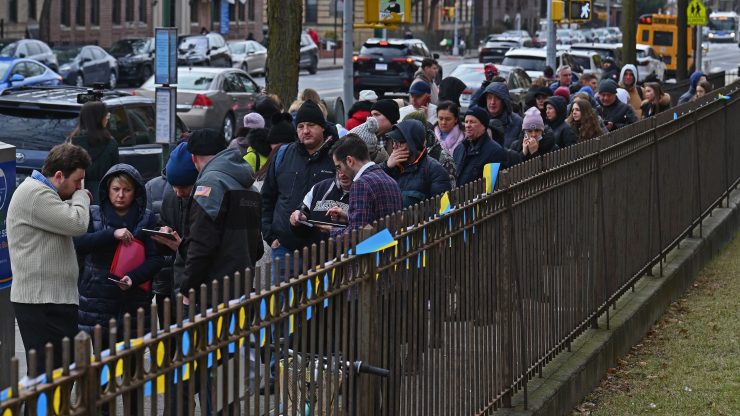 Ukrainian refugees stand in line to attend a job fair in Brooklyn on Feb. 1. It is cold, so people are wearing jackets. They line up along a fence that has small Ukrainian flags on it.