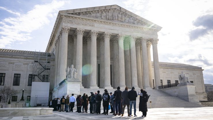 A crowd of people gather in line outside of the U.S. Supreme Court, a large, traditional structure with steps leading to a portico supported by eight columns. It is a partly cloudy day, and the sun is seen behind the Court building. A ray of light streaks across the building.