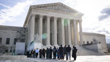 A crowd of people gather in line outside of the U.S. Supreme Court, a large, traditional structure with steps leading to a portico supported by eight columns. It is a partly cloudy day, and the sun is seen behind the Court building. A ray of light streaks across the building.