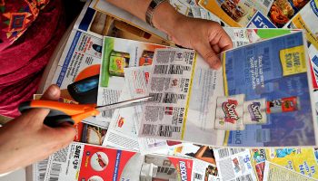 A person uses orange-handled scissors to cut coupons from an array of printed inserts.