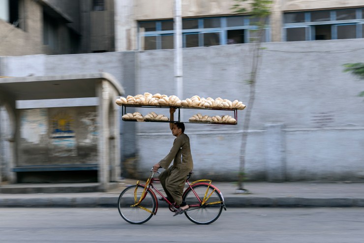A worker wearing brown clothing rides a bicycle, the road and urban surroundings by him blurred by his motion. One hand is on the bike's steering wheel, the other holds a long rack that holds two shelves of bread, which is balanced on his head.