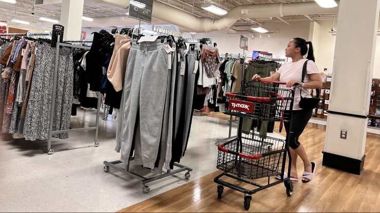 A woman pushes a cart past a rack of gray pants in a T.J.Maxx. Off to the side are long racks of clothes.