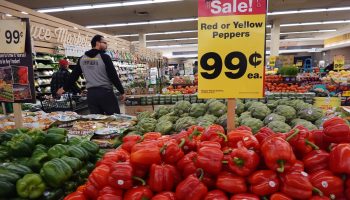 A sign reads "Sale: Red or Yellow Peppers" in a produce aisle at a grocery store. Shoppers mill about in the background.