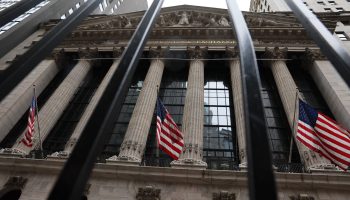 The exterior of the New York Stock exchange, a portico supported by stone columns, is seen through gate bars. Several American flags are raised outside of the building.