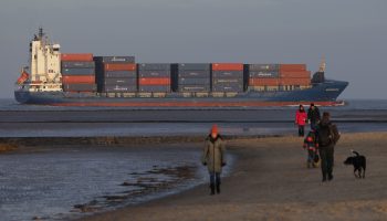 Winter beachgoers stroll along the beach as a container ship transports goods in the background.