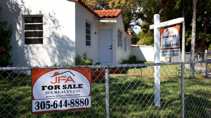 Two for sale signs — one hanging from a white post, the other posted on a grey chainlink fence — are seen in the green grassy yard of a single-story, Spanish-style home, with white stucco and red clay roof shingles.