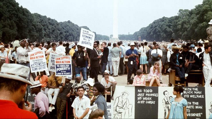 Hundreds of people gather at the end of the Poor People March on June 19, 1968, in Washington DC. They hold signs advocating the end of poverty. The Washington Monument is seen on the other side of the mall.