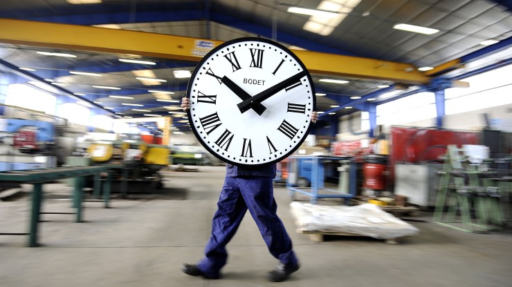 A worker carries a big clock face in front of their body.
