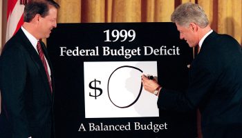 Vice President Al Gore watches President Bill Clinton place a zero next to a dollar sign on a board reading: 1999 Federal Budget Deficit. Underneath the $0 are the words: A Balanced Budget.