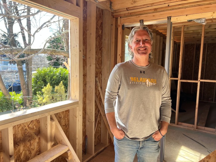 Matt Hampton, a man in a gray long sleeve shirt and blue jeans with gray hair, stands in the new addition under construction at his house. He is surrounded by wood wall framings with trees viewable in a spot for a window.