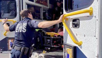 A man in a blue uniform with white lettering on the back reading "New Orleans EMS" faces the inside of an ambulance patient compartment, his arms stretched across the opening and hands holding yellow rails. Inside, another worker stands over a gurney.