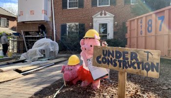 A wooden sign outside a brick, two-story home reads "Under Construction." In the background are a framed and sheeted addition, an orange dumpster and two stuffed pigs wearing construction hats.