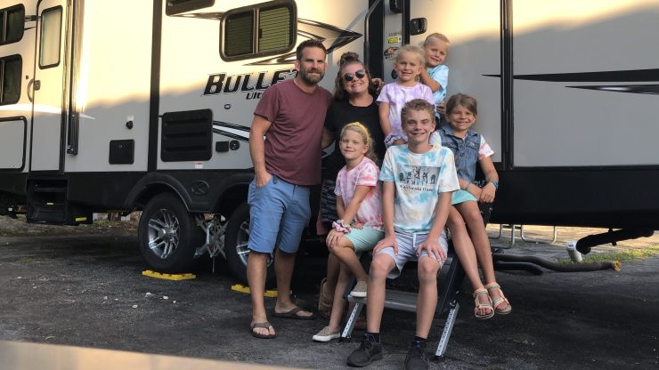 Brian and Shannon Ostrovsky stand with their five children, posing for a picture outside of a travel trailer.