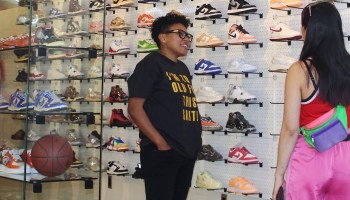 Venita Cooper, owner of Silhouette Sneakers & Art, wears all black and stands in front of a wall holding shelves lined with dozens of single multicolored sneakers. The back of a customer with long black hair, a red tank top and pink shorts, is seen, as they are looking at Venita.