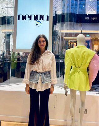 Yana Smaglo stands in front of a storefront display window with a mannequin to her side. She is wearing blackpants, with a denim shorts accessory and white shirt. The mannequin behind her, facing out, wears a neon jacket with one poofy pink sleeve. Behinder Smaglo is a light up sign that says "Nenya."