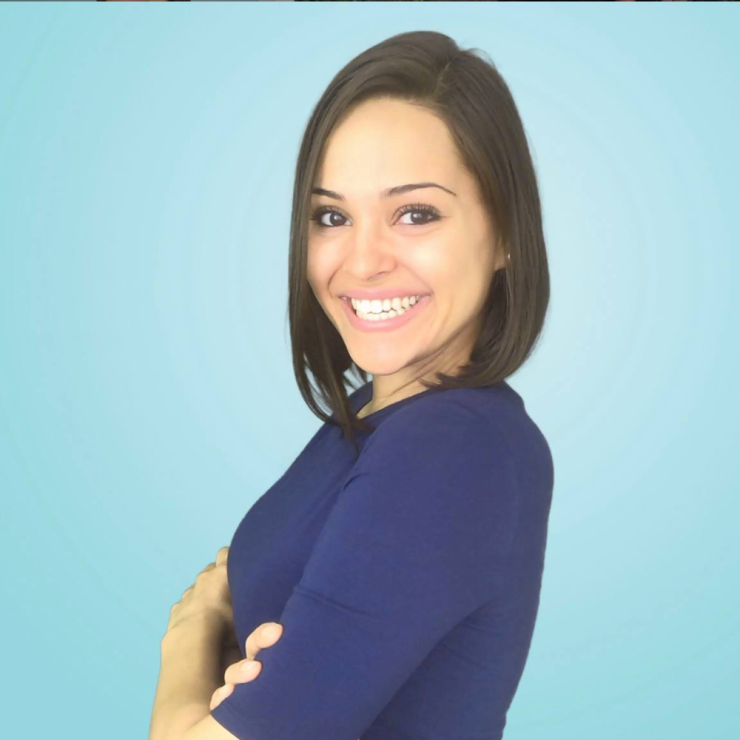 Yanely Espinal has shoulder-length brown hair and a dark blue dress.  She is standing in front of a light blue background, smiling with her arms crossed over her cheek.
