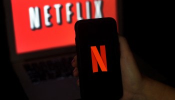 The Netflix logo on a screen: a red field with white letters outlined in black. In front of the screen is a cellphone with a red N on a black field, a smaller version of the logo.