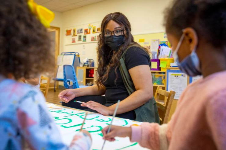A woman with long, dark hair, a black short sleeve shirt and army green vest sits at a children's table in an early learning center. She is looking forward at two children who are blurred by the camera. They are painting letters in bright green.