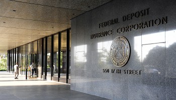 A front view of an FDIC building, with people entering the floor-ceiling mirrored glass doors. To the right are panels of stationary glass and then a polished stone wall with the words: Federal Deposit Insurance Corp., 550 17th Street. Between the name and address is the FDIS seal.