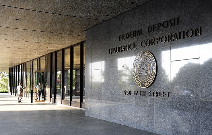 A front view of an FDIC building, with people entering the floor-ceiling mirrored glass doors. To the right are panels of stationary glass and then a polished stone wall with the words: Federal Deposit Insurance Corp., 550 17th Street. Between the name and address is the FDIS seal.