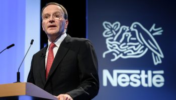 Nestle CEO Mark Schneider, a man wearing glasses, a black suit, white shirt and red tie, stands and speaks at a dark wood lectern in front of a white Nestle logo.