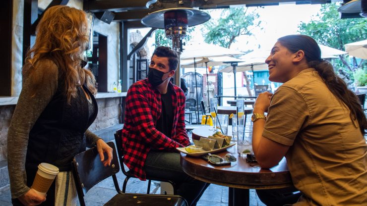 Two people, one with short black hair, a black face mask and red plaid shirt, the other with long dark hair and a gray shirt, sit at a high-top table on a restaurant's outdoor patio. They smile and look at a waitress with long sandy blonde hair and a black shirt.