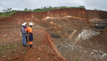 Three workers in white hard hats and work gear look out over an open cast lithium mine. It is a a great gorge with rust, brown and white colored rocks. At the sides of the massive hole, greenery grows.