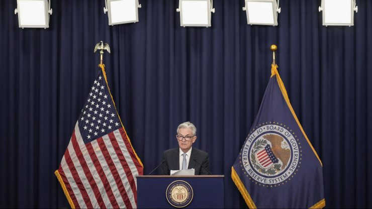 Fed Chair Jerome Powell, an older man with gray hair, a black suit, white shirt, purple tie and black glasses, stands at a podium. He is giving remarks under a series of white overhead lights, in front of blue drapery, and an American flag on the left and Federal Reserve flag on the right.