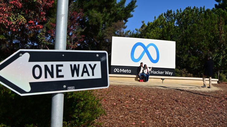 Two people smile and pose for a photo near the Meta logo sign, which shows a blue infinity symbol shaped like an M. It is located outside and trees are shown behind mulch, in front of trees, and behind a one-way roadsign.