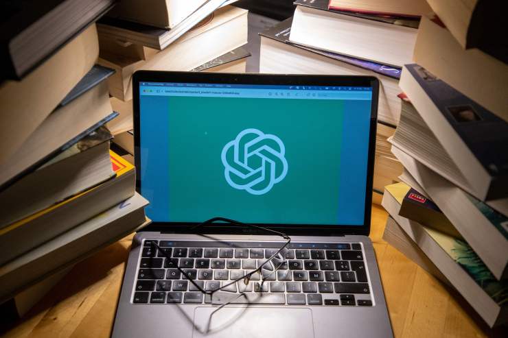 An open laptop with the ChatGPT logo on the screen — sort of a hexagon with rounded edges and interlinking arms. Stacks of books surround the laptop.