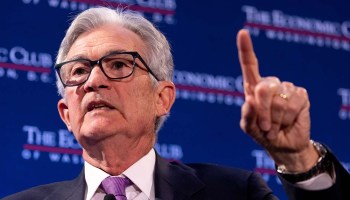 Federal Reserve Board Chairman Jerome Powell, an older man with gray hair, black frame glasses, a white shirt, purple tie and black suit, is talking with his left index finger held up.