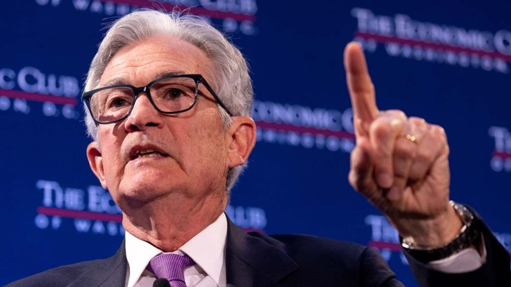 Federal Reserve Board Chairman Jerome Powell, an older man with gray hair, black frame glasses, a white shirt, purple tie and black suit, is talking with his left index finger held up.