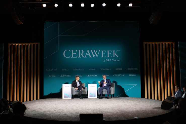 Two men in dark suits sit on a stage underneath a sign that reads "CERAWEEK by S&P Global." The silhouettes of audience members' heads are in the foreground.