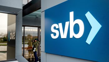 A person is seen pointing a camera at a building. In front of him is a large gray pillar, with a blue sign displaying the SVB logo. (Lower-case "svb" in a white, sans serif text with a blue caret.