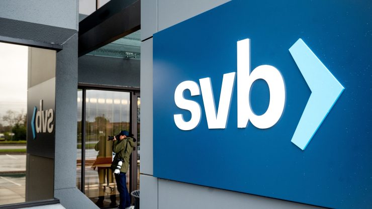 A person is seen pointing a camera at a building. In front of him is a large gray pillar, with a blue sign displaying the SVB logo. (Lower-case "svb" in a white, sans serif text with a blue caret.