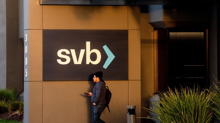 A man wearing a backpack and holding a cellphone passes under a sign reading "svb›" in front of Silicon Valley Bank's headquarters in Santa Clara, California.
