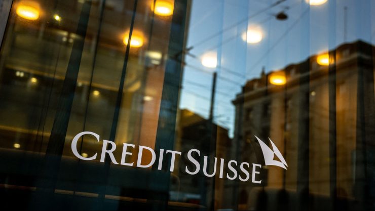 The white logo for Credit Suisse is seen on a glass window pane. A series of warm overhead lights are seen within.