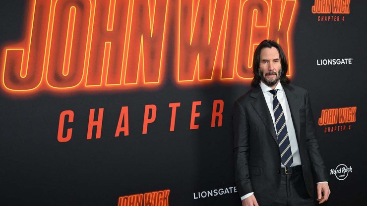 Keanu Reeves, a man with long black hair and a beard wears a black suit, with a white shirt and black and white striped tie. He stands in front of a promotional for his film "John Wick: Chapter 4."