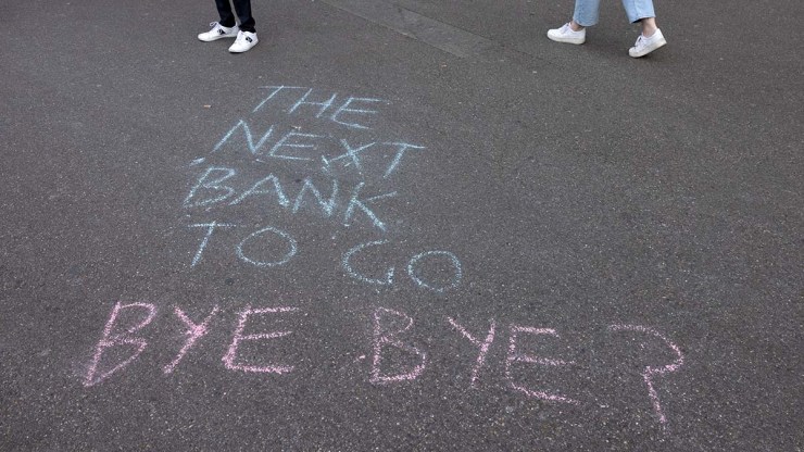 Blue and pink chalk spell out "The Next Bank To Go Bye Bye?" on concrete while two pedestrians walk by.