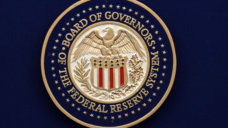 At the center of the round seal of the Federal Reserve Board of Governors is a spread-winged bald eagle sitting atop a shield with stars across the top and red and white vertical stripes below the stars.