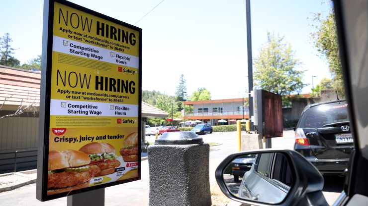 A "Now Hiring" sign is displayed on a digital screen in a McDonald's drive-thru.