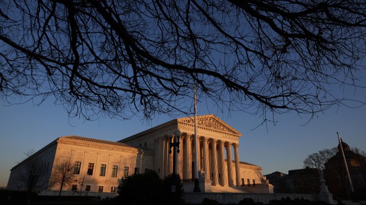 The U.S. Supreme Court building is seen in warm sunlight and is set against a clear blue sky. Branches of a tree come down above the Court in the foreground.