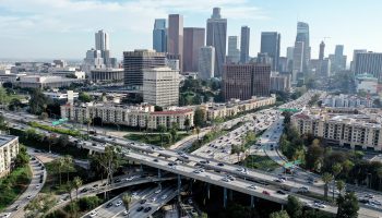 The skyline of downtown Los Angeles is seen behind highways filled with dozens of moving vehicles. Some greenery is seen and the skyline of apartments and office buildings is set against a partly cloudy sky.