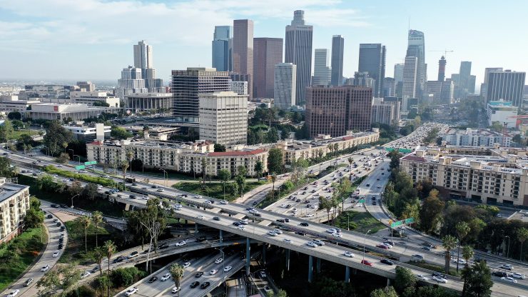 The skyline of downtown Los Angeles is seen behind highways filled with dozens of moving vehicles. Some greenery is seen and the skyline of apartments and office buildings is set against a partly cloudy sky.