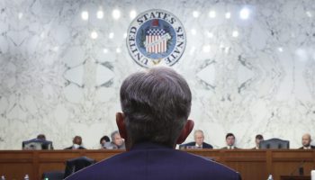 The back of Fed Chair Jerome Powell, with a blue blazer and head of grey hair, is seen before a long wood panel at which members of the Senate Banking, Housing, and Urban Affairs Committee are seated. Behind them is a white marble wall reflecting overhead lights and the seal of the Senate.